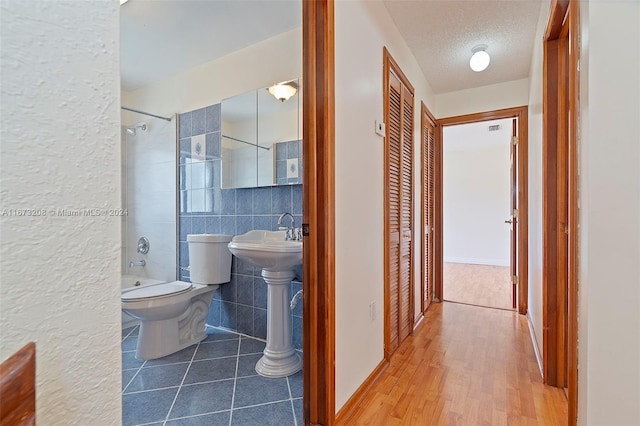 bathroom with a textured ceiling, hardwood / wood-style floors, toilet, tile walls, and tiled shower / bath combo