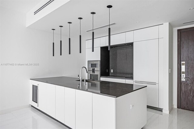 kitchen with a center island with sink, sink, hanging light fixtures, stainless steel double oven, and white cabinetry