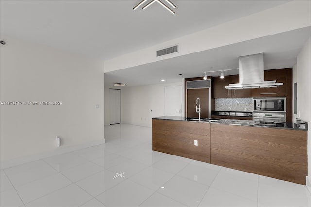 kitchen featuring decorative backsplash, exhaust hood, light tile patterned floors, built in appliances, and dark brown cabinetry