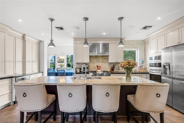 kitchen with a healthy amount of sunlight, stainless steel appliances, wall chimney range hood, and a kitchen island with sink