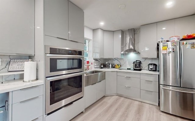 kitchen featuring wall chimney exhaust hood, stainless steel appliances, sink, light wood-type flooring, and tasteful backsplash