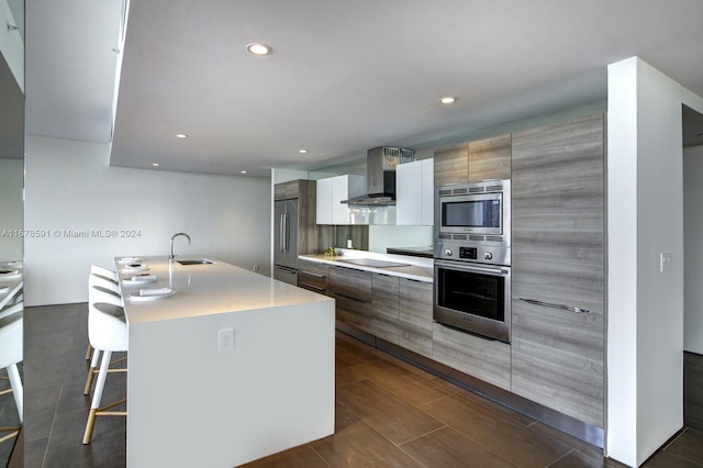 kitchen with wall chimney range hood, appliances with stainless steel finishes, sink, a kitchen breakfast bar, and a kitchen island with sink