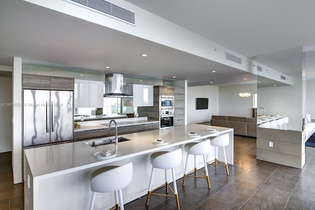 kitchen with wall chimney range hood, sink, built in appliances, and a kitchen breakfast bar