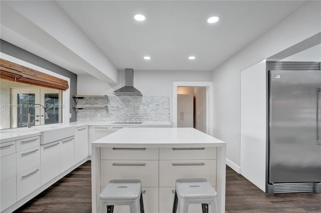 kitchen with wall chimney range hood, open shelves, modern cabinets, stainless steel built in refrigerator, and dark wood-type flooring