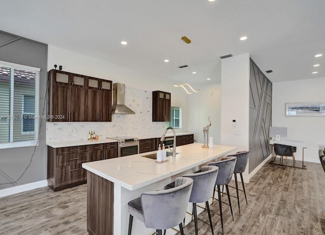 kitchen featuring stainless steel electric stove, an island with sink, light hardwood / wood-style floors, wall chimney exhaust hood, and sink