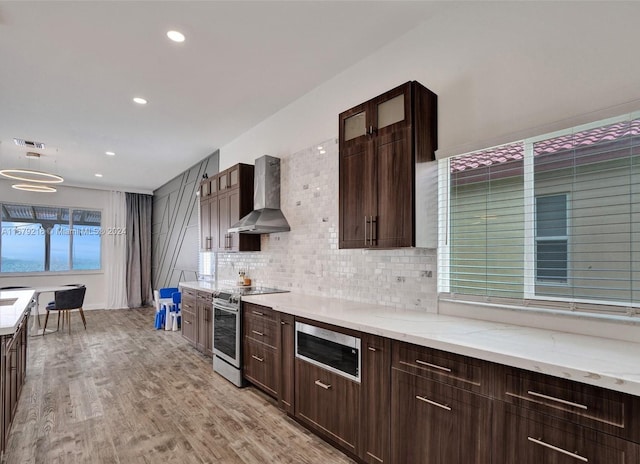 kitchen featuring tasteful backsplash, light stone countertops, appliances with stainless steel finishes, light hardwood / wood-style floors, and wall chimney exhaust hood