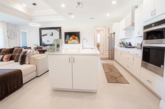 kitchen with stainless steel appliances, white cabinets, light countertops, wall chimney exhaust hood, and an island with sink