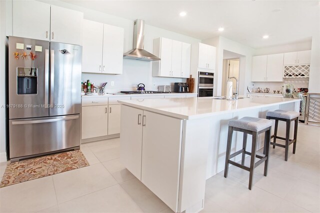 kitchen with wall chimney exhaust hood, white cabinets, stainless steel appliances, and an island with sink