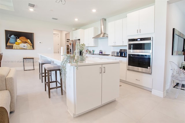 kitchen with wall chimney exhaust hood, a kitchen breakfast bar, a center island with sink, white cabinetry, and appliances with stainless steel finishes