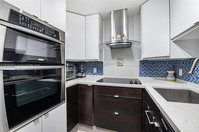 kitchen featuring stainless steel double oven, white cabinets, a sink, wall chimney range hood, and black electric cooktop