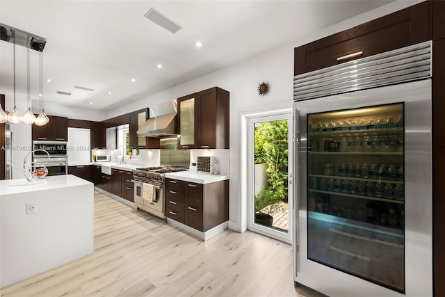 kitchen with wall chimney range hood, beverage cooler, appliances with stainless steel finishes, light hardwood / wood-style floors, and decorative light fixtures