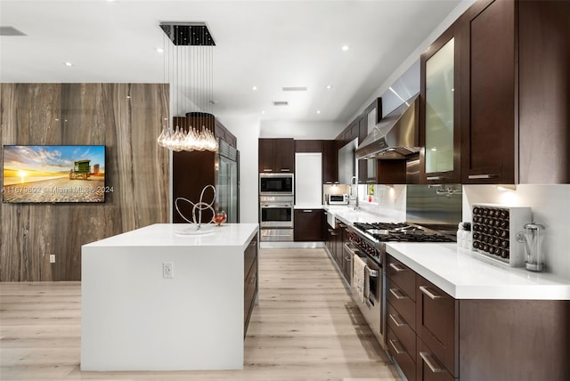 kitchen with appliances with stainless steel finishes, wall chimney range hood, a center island with sink, and light wood-type flooring