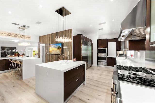 kitchen with a kitchen island, light hardwood / wood-style floors, wall chimney exhaust hood, pendant lighting, and built in appliances