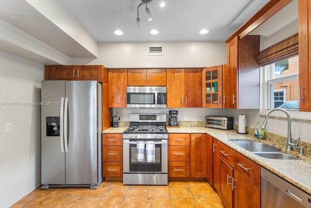 kitchen with light stone counters, appliances with stainless steel finishes, brown cabinetry, glass insert cabinets, and a sink