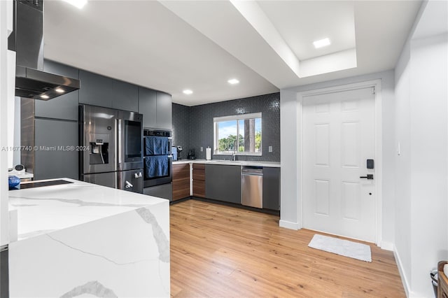 kitchen featuring wall chimney range hood, sink, light stone countertops, appliances with stainless steel finishes, and light hardwood / wood-style floors