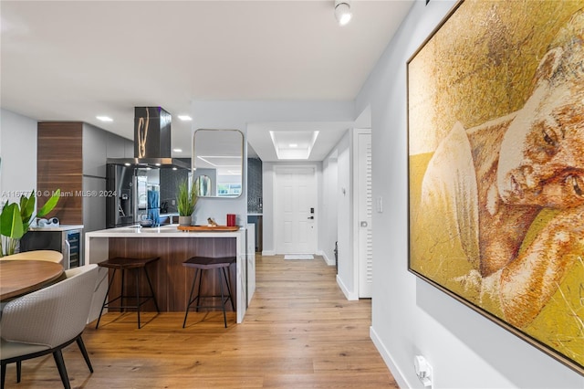 kitchen with light wood-type flooring, wall chimney range hood, kitchen peninsula, and a kitchen bar