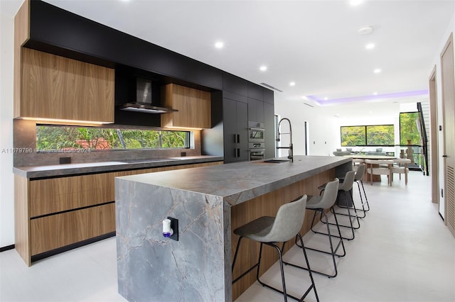 kitchen with sink, wall chimney exhaust hood, stainless steel double oven, a kitchen island with sink, and a breakfast bar