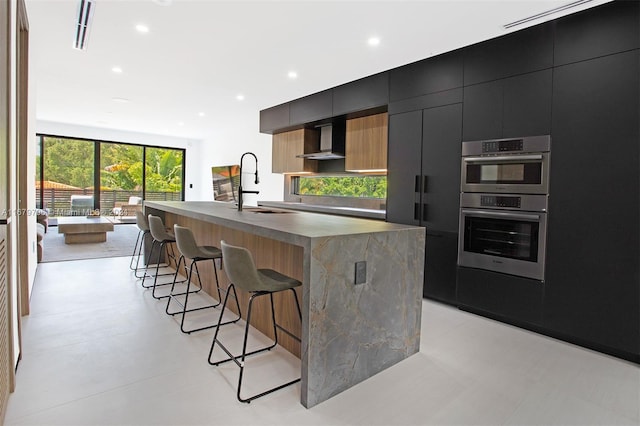 kitchen with sink, stainless steel double oven, wall chimney range hood, a kitchen breakfast bar, and an island with sink