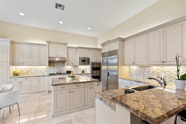 kitchen featuring built in appliances, sink, backsplash, a kitchen island with sink, and light stone counters