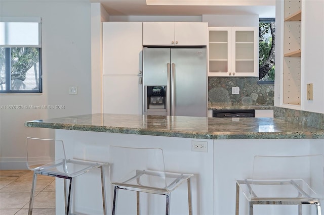 kitchen with kitchen peninsula, white cabinetry, stainless steel fridge with ice dispenser, decorative backsplash, and light tile patterned floors