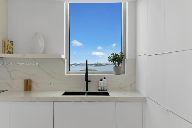 kitchen featuring a water view, backsplash, white cabinets, a sink, and light stone countertops