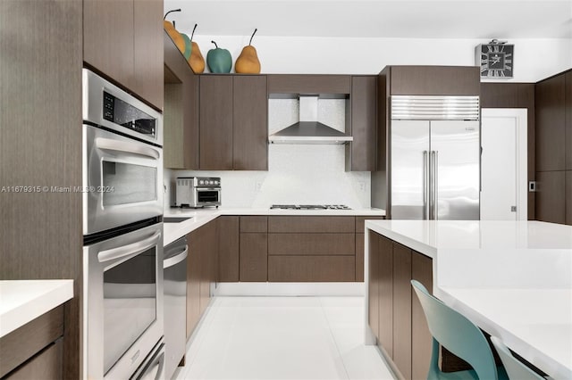 kitchen with appliances with stainless steel finishes, wall chimney exhaust hood, dark brown cabinets, and light tile patterned floors