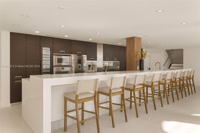 kitchen featuring built in appliances, a large island, dark brown cabinetry, and a breakfast bar