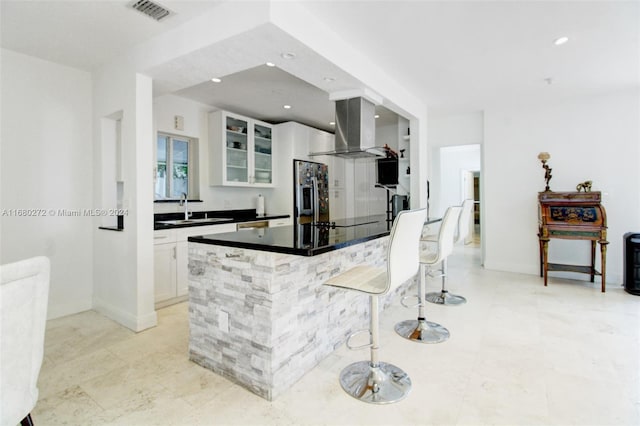 kitchen with wall chimney range hood, white cabinets, stainless steel fridge with ice dispenser, a breakfast bar, and sink