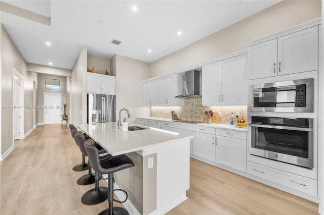 kitchen with stainless steel appliances, sink, white cabinets, wall chimney exhaust hood, and a kitchen island with sink