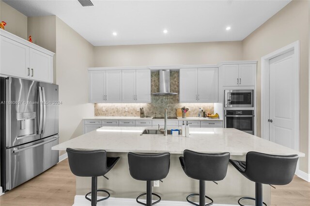 kitchen featuring an island with sink, wall chimney range hood, white cabinetry, and appliances with stainless steel finishes