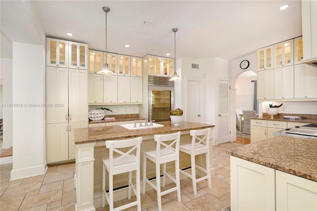 kitchen featuring stone counters, sink, stainless steel built in refrigerator, hanging light fixtures, and a center island with sink