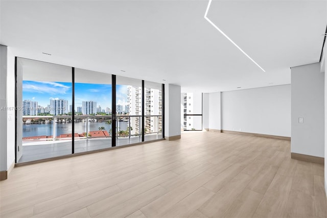 empty room featuring a wall of windows, baseboards, a water view, a view of city, and light wood-type flooring