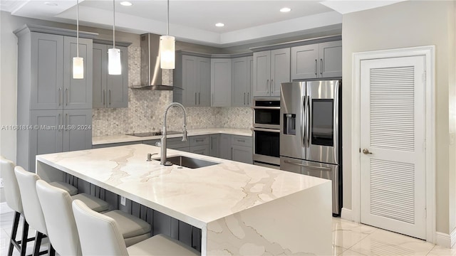 kitchen featuring stainless steel appliances, light stone countertops, wall chimney exhaust hood, decorative light fixtures, and an island with sink