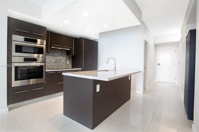 kitchen with decorative backsplash, stainless steel double oven, a center island with sink, sink, and light tile patterned floors