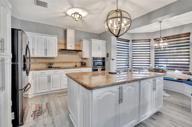 kitchen featuring a kitchen island with sink, wall chimney range hood, pendant lighting, white cabinetry, and appliances with stainless steel finishes