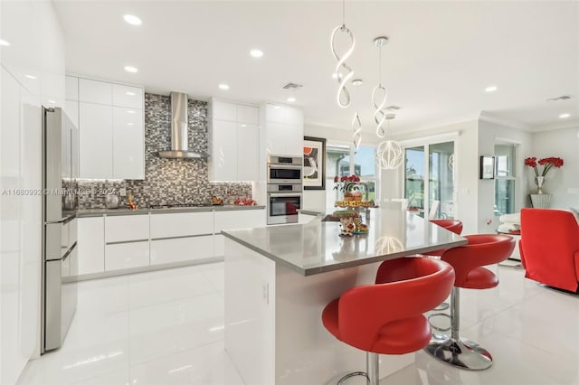 kitchen featuring a kitchen island, wall chimney range hood, a breakfast bar area, decorative light fixtures, and white cabinetry