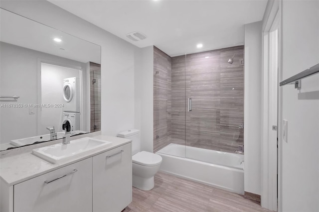 full bathroom with hardwood / wood-style floors, stacked washing maching and dryer, shower / bath combination with glass door, toilet, and vanity