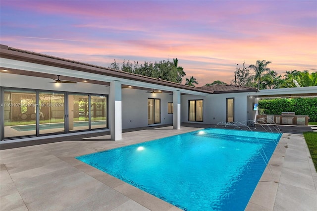 pool at dusk with a patio, exterior kitchen, pool water feature, and ceiling fan