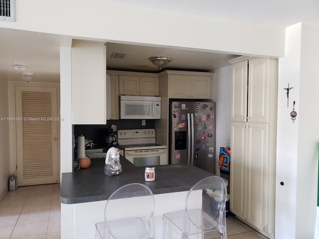 kitchen featuring white appliances, light tile patterned floors, cream cabinets, kitchen peninsula, and a breakfast bar