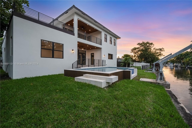 back house at dusk featuring a lawn, ceiling fan, a swimming pool with hot tub, and a balcony