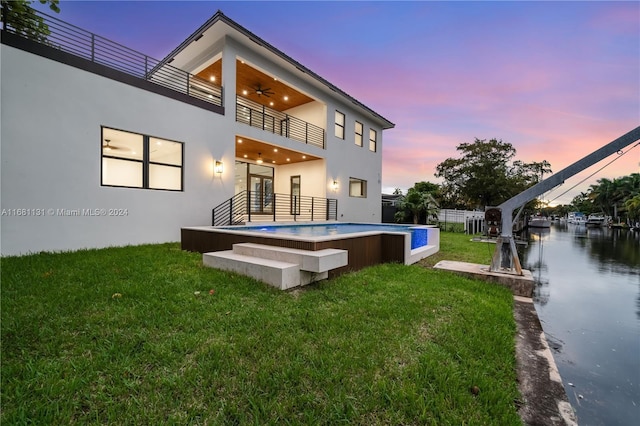 back house at dusk with ceiling fan, a balcony, a water view, and a lawn