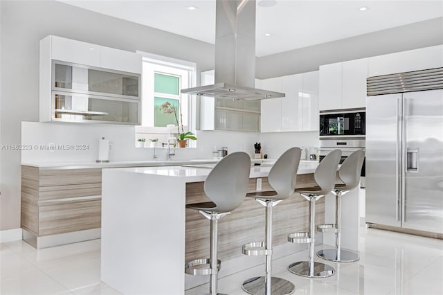 kitchen featuring white cabinets, a breakfast bar, island range hood, built in appliances, and a center island