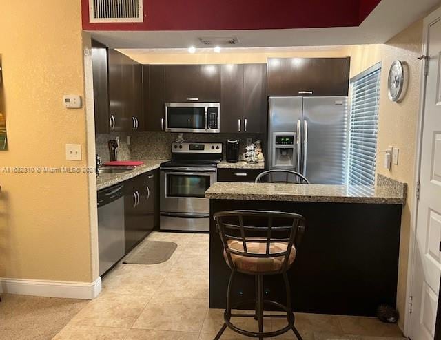 kitchen featuring decorative backsplash, dark brown cabinets, light stone countertops, a kitchen bar, and appliances with stainless steel finishes