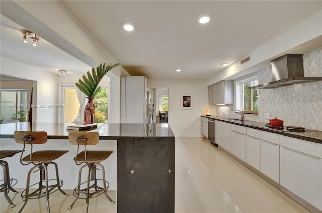 kitchen featuring decorative backsplash, a sink, wall chimney range hood, stovetop, and a kitchen bar