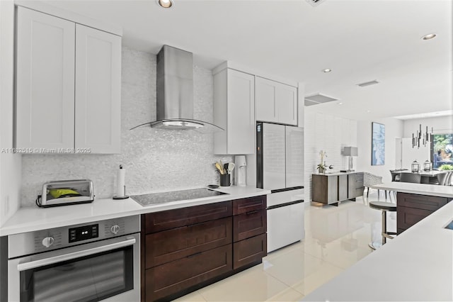 kitchen with stainless steel oven, black electric stovetop, wall chimney exhaust hood, white fridge, and white cabinetry