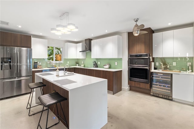 kitchen with beverage cooler, light countertops, appliances with stainless steel finishes, wall chimney range hood, and an island with sink