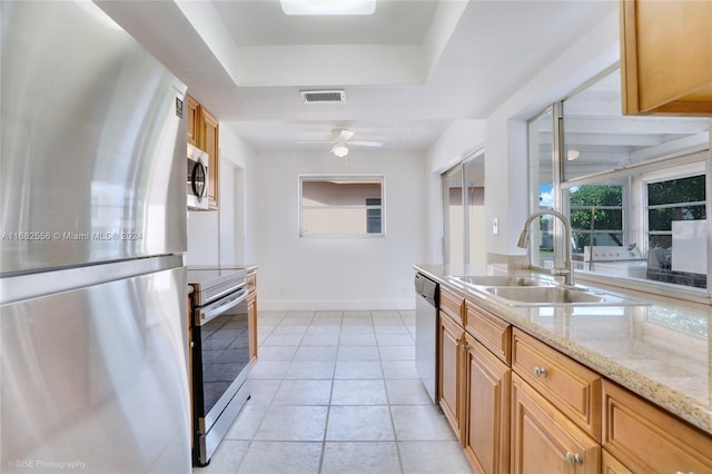 kitchen with a raised ceiling, appliances with stainless steel finishes, ceiling fan, light stone countertops, and sink