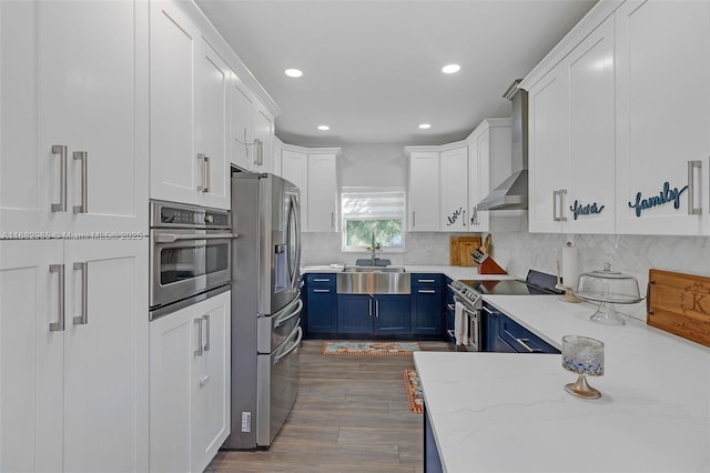 kitchen with blue cabinets, white cabinetry, light stone counters, stainless steel appliances, and wall chimney range hood