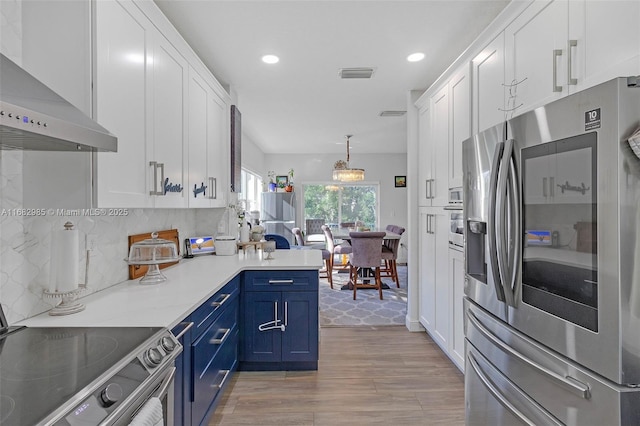 kitchen with appliances with stainless steel finishes, white cabinets, hanging light fixtures, blue cabinetry, and wall chimney range hood