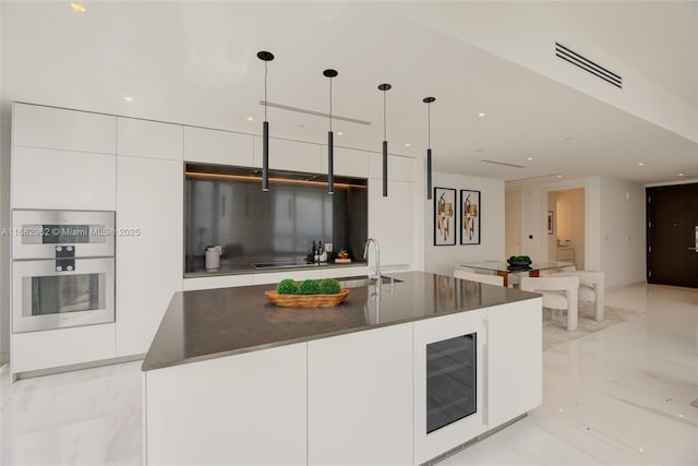 kitchen with white cabinets, decorative light fixtures, wine cooler, and an island with sink